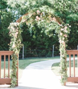 Wooden wedding arch draped in greenery and pale pink flowers on a sunny outdoor path.