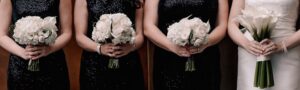 Three bridesmaids in black dresses hold white rose and hydrangea bouquets, with the bride in white on the right holding a white bouquet of lilies separate from them.