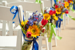 Bouquet of sunflowers, red tulips, and purple flowers tied to white chairs with blue ribbons along an aisle.
