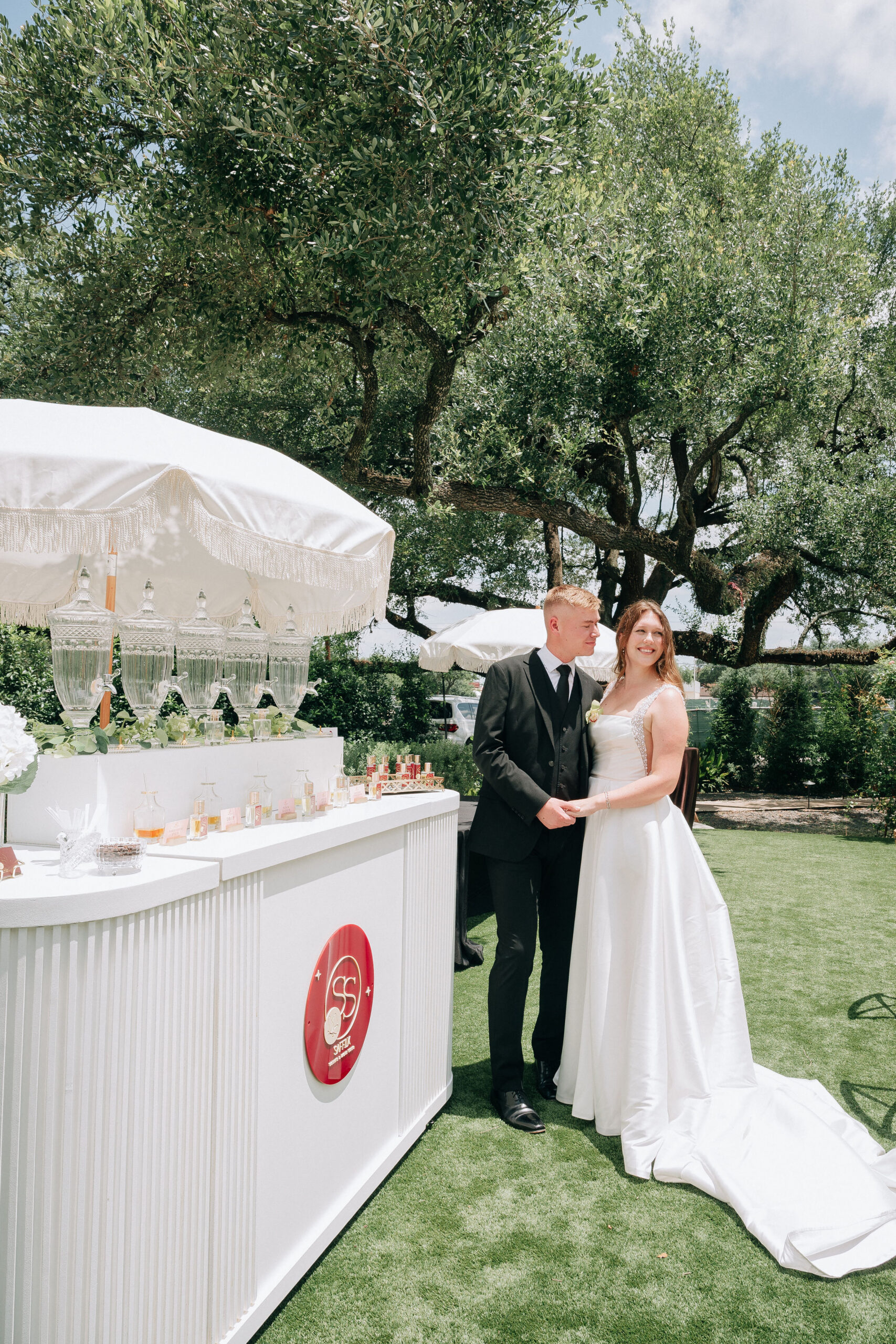 Bride and groom in formal wear stand by a white outdoor drink bar with crystal dispensers under trees.