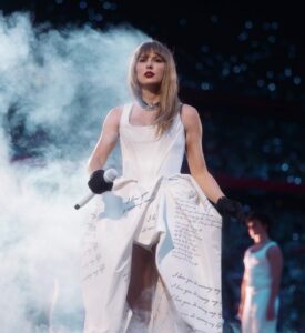 Female stage performer in a white dress with handwritten-text skirt, holding a microphone amid blue smoke on a dimly lit stage.
