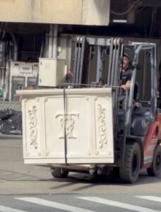 Forklift carrying a large white decorative cabinet with carved scrollwork in an industrial yard