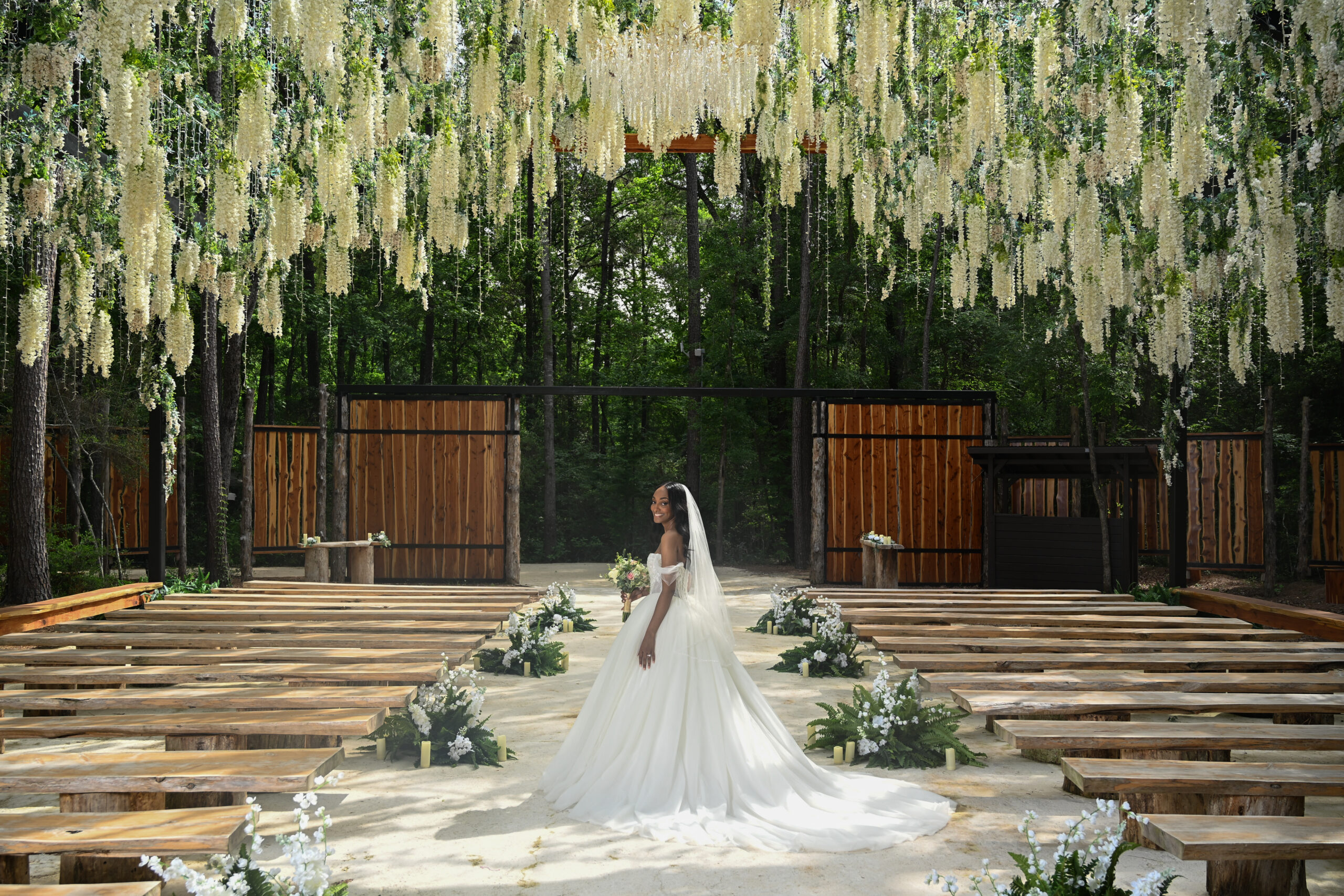 Bride in a white wedding gown walks down an outdoor aisle under hanging white flowers, with wooden benches and greenery nearby.